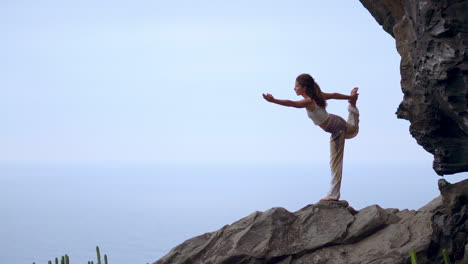 Amid-the-tranquil-blue-ocean-scenery-at-sunset,-a-young-woman-practices-yoga-on-a-rocky-seashore,-illustrating-a-healthy-lifestyle,-harmony,-and-the-seamless-relationship-between-humans-and-nature
